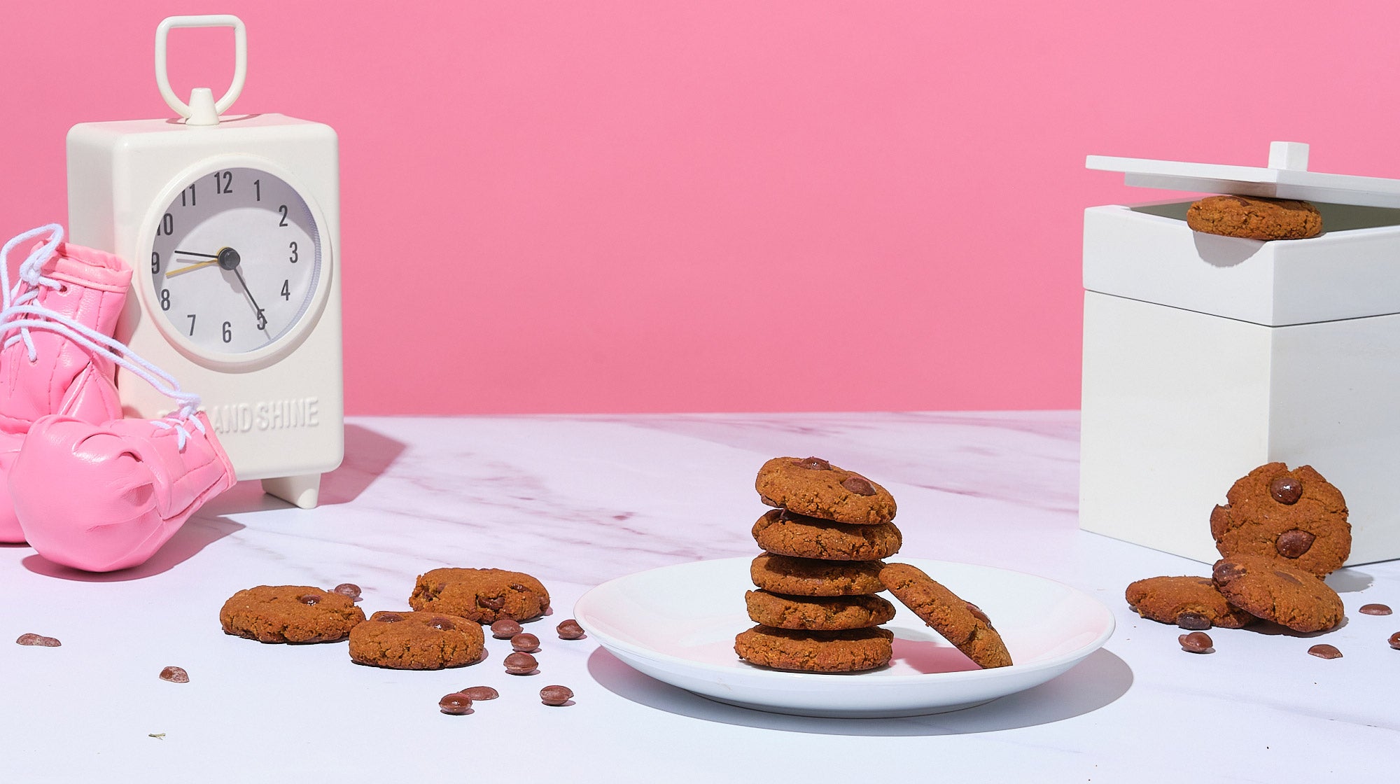 Bake Me Healthy Gluten-free, vegan, dark chocolate chip cookies stacked on  a plate next to cookie box and clock with boxing gloves